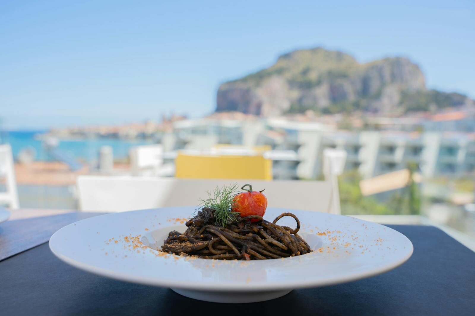 Plate of squid ink pasta with tomato and herbs, set against a beautiful seaside view.