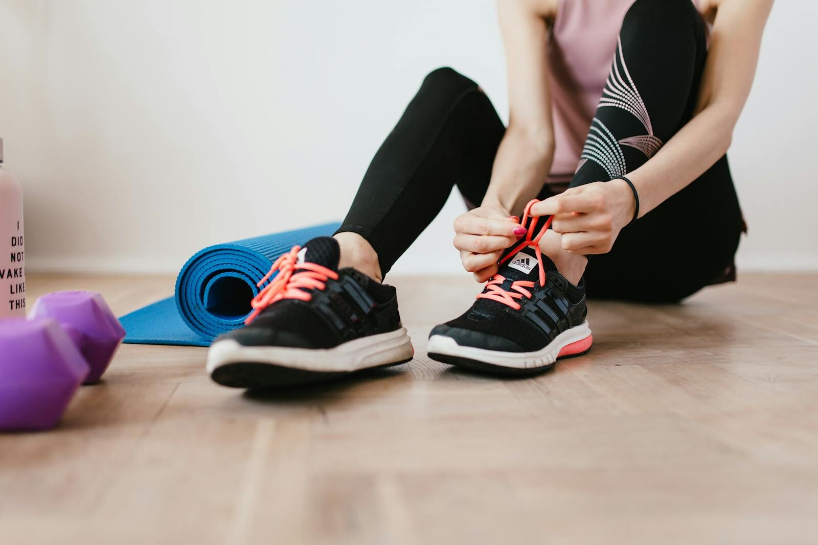 Crop sportswoman in sportswear tying sneakers while sitting on wooden floor with sport mat and dumbbells before exercising indoors