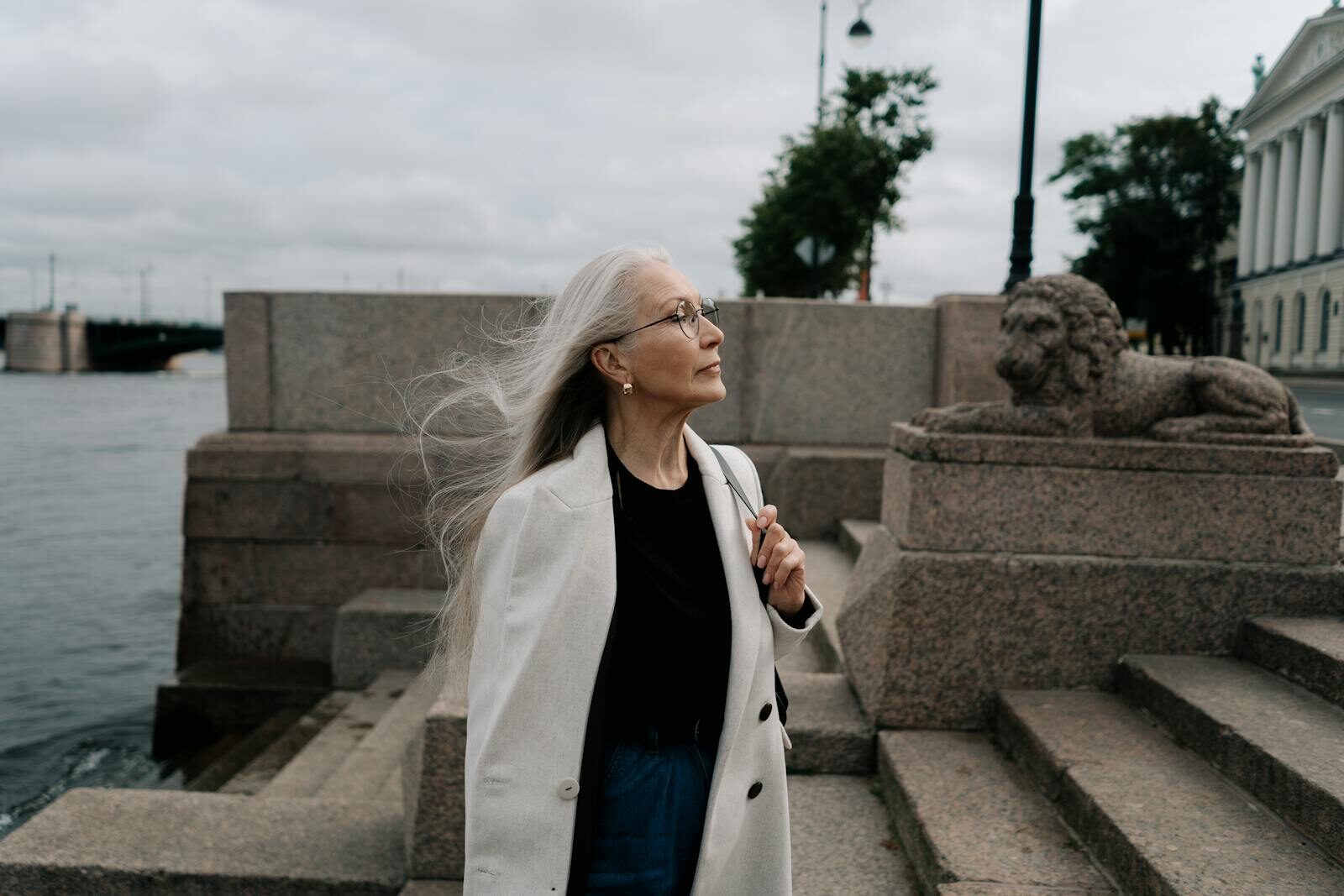 Senior woman with gray hair and glasses in white blazer outdoors near river and stone lion statue.