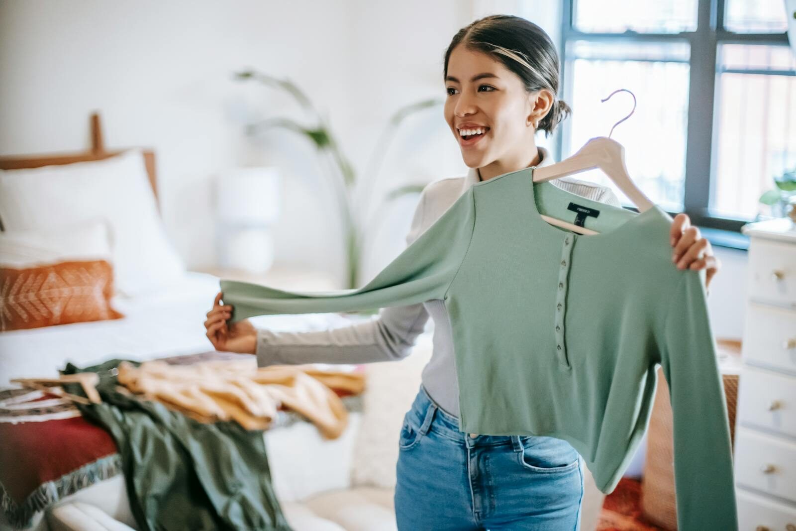 Positive ethnic woman in trendy outfit selecting clothes while demonstrating blue blouse in light room at home and looking away